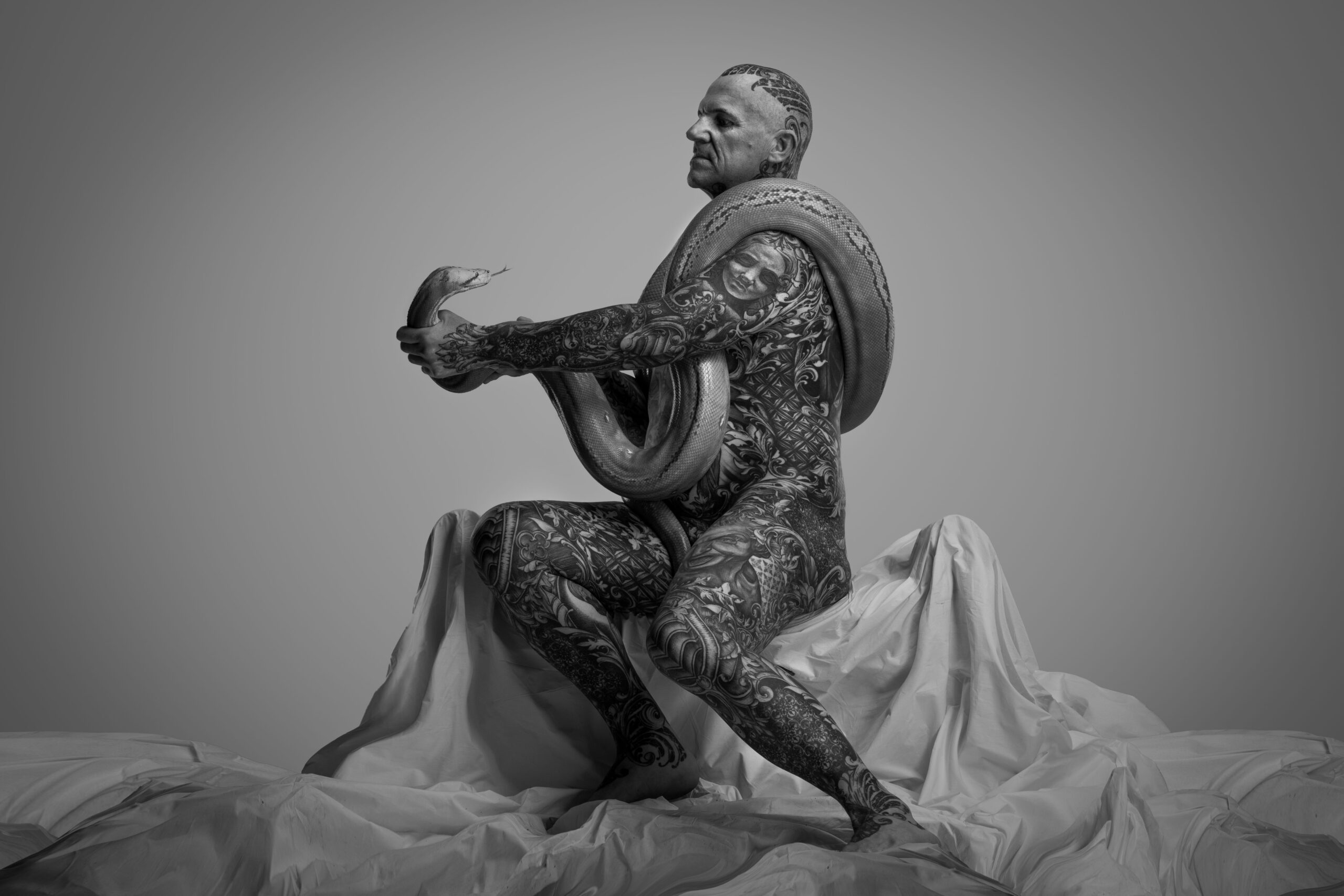 serpente-emozioni-sulla-pelle-spazioq Lee Robert Ness fotografato da Silvia Qadrini che tiene a distanza con le proprie mani un boa attorvigliato al proprio corpo e lo guarda negli occhi